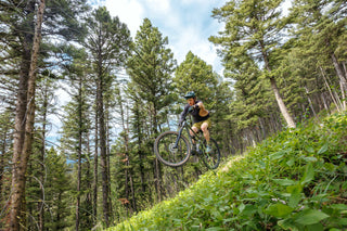 Chad boosts of a kicker on his mountain bike wearing the swamp king kit over a field of green somewhere in the Bridgers Mountain Range in Montana