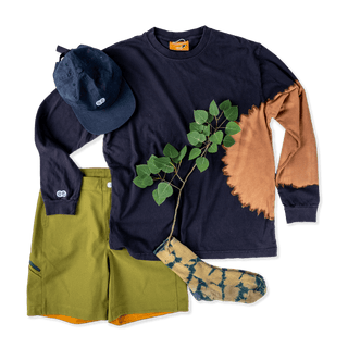 Lifestyle lay down of black longsleeve coupled with avacado green mtb shorts. An Aspen tree branch lays across it all with a hat and socks.