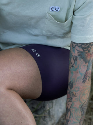 Close up of holly sitting on a log wearing her light green pocket tee and dark purple bike camois with laser cut hem