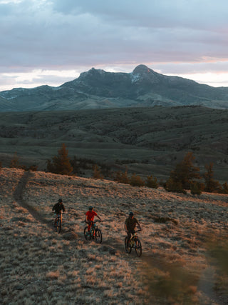 Three mountain bikers ride along a dirt trail across a grassy hillside, with rolling hills and a large mountain range under a cloudy sky in Cody Wyoming.