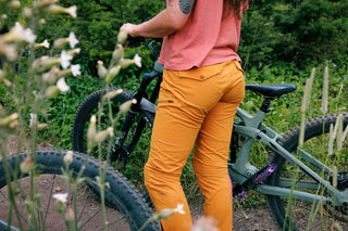 Lila walks her bike sporting the sleek and stylish buckthorn brown pants with rear flap pockets and matching snaps