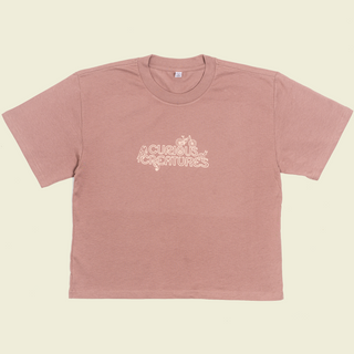 Curious Creatures logo print on the front of a hazy pink crop top tee shirt outlined with bugs and bikes