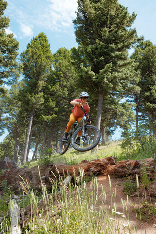 Lila boosts off a fallen log on a mountain bike trial wearing her pocket tee and pants on a blue bird day