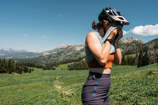 Hannah wipes sweat from her face using her Steel Blue Tank atop red cub trail with a breath taking Montana view, showing off her high waisted purple shorts