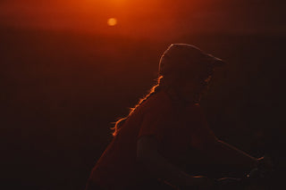 A person riding a bicycle in silhouette against a dramatic orange sunset sky, with the sun low on the horizon and strong backlighting.
