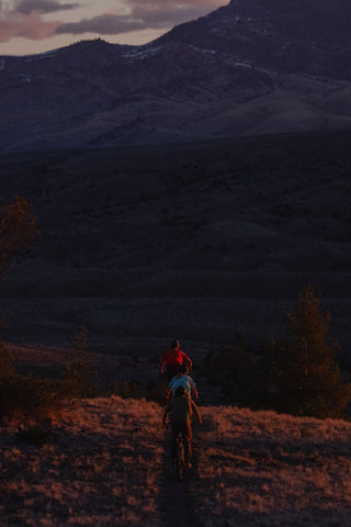 A group of three rides bikes along a dirt trail in a mountainous outdoor landscape at dusk, with dark purple hills and mountains in the background.