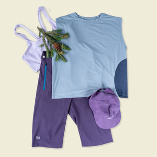 Lifestyle lay down of blue tank with purple shorts along with a purple sports bra and purple hat topped with a pine branch