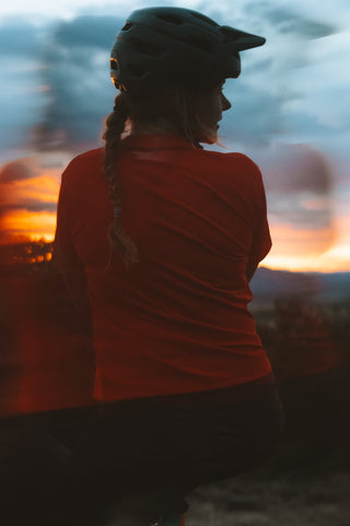 A girl seen from behind wearing a bicycle helmet and a bright red short-sleeve jersey, sitting outdoors against a dramatic sunset sky with blurred orange and blue tones.