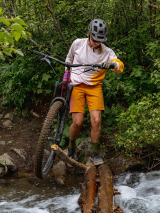 Hillary crosses a roaring creek across some slippery logs with her bike in hand