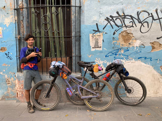 Pasha leans up against an old building with a couple of beers and bikes in the city of Oaxaca in Mexico after a long bike pack adventure