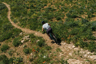 Hannah shreds downhill on her mountain bike across a narrow strip of dusty rocks through wildflowers