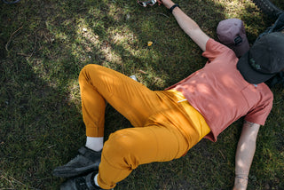 Hannah sprawls out on the shady grass with hat over her head wearing a pink pocket tee and mustard pants